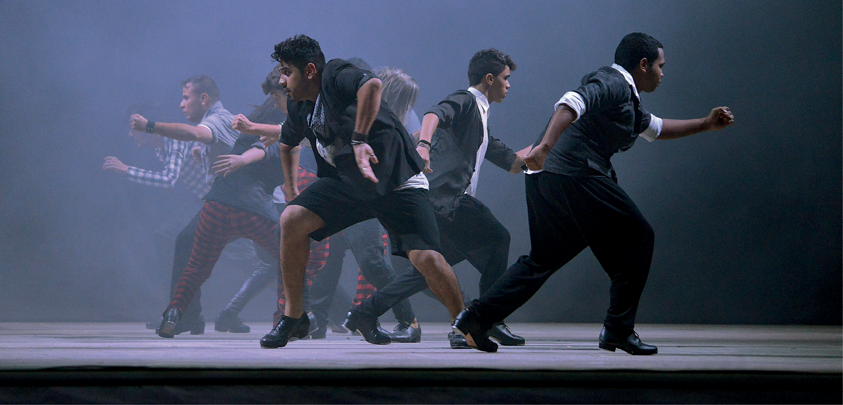 Fotografia. Homens dançam em um palco com fundo enevoado, voltados para lados opostos. Alguns vestem jaqueta, camisa e calça pretas. Um deles veste bermuda preta.
