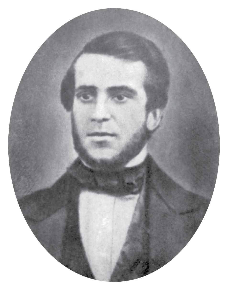 Fotografia em preto e branco. Retrato de um homem de cabelo escuro e sobrancelhas espessas, com barba em torno do queixo. Ele usa gravata-borboleta, camisa clara e casaco escuro.