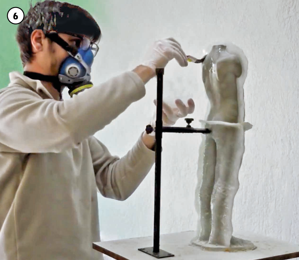 6: O artista, vestido com blusão bege, usa máscara de proteção e luvas de látex e aplica, com um pincel, um produto químico transparente sobre o corpo da escultura, que está sem os braços e a cabeça, apoiada em um suporte, sobre uma pequena bancada.