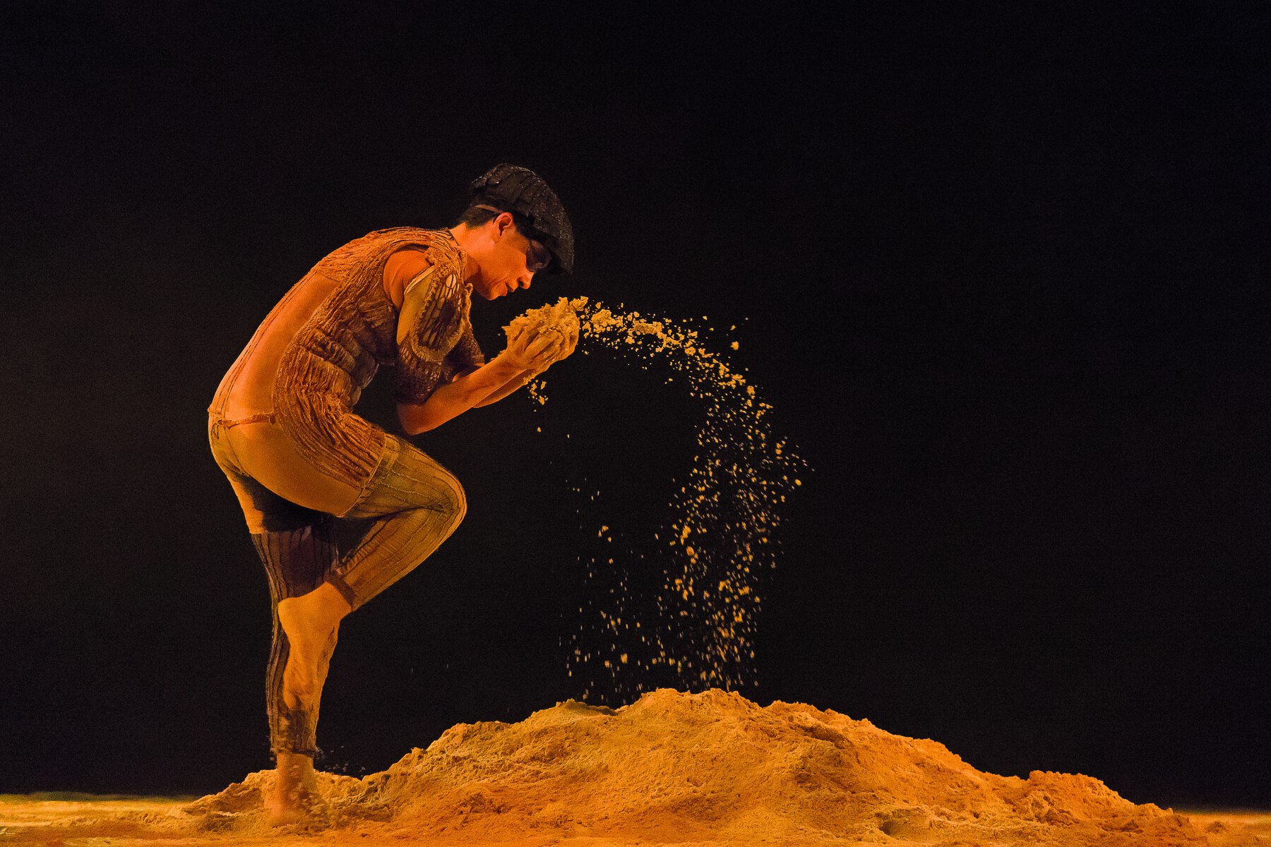 Fotografia. Dançarina executa movimento de dança no palco. Ela usa uma boina e veste uma roupa justa em tons de marrom, com remendos. Seu corpo estás levemente curvado para frente, a perna direita está dobrada e ela lança um punhado de terra com as mãos. No chão, um monte de terra.