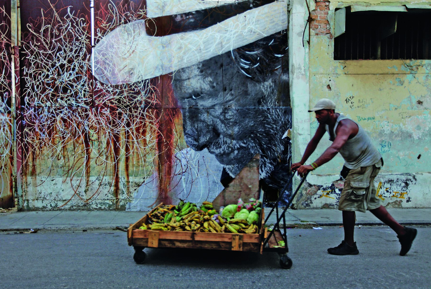 Fotografia. Em primeiro plano, um homem de boné, camiseta regata e bermuda empurra um carrinho de mão com muitas frutas. Ao fundo, muro com grafite do perfil do rosto de um homem idoso com chapéu, barba e bigode brancos. Ele está com a cabeça levemente voltada para baixo.