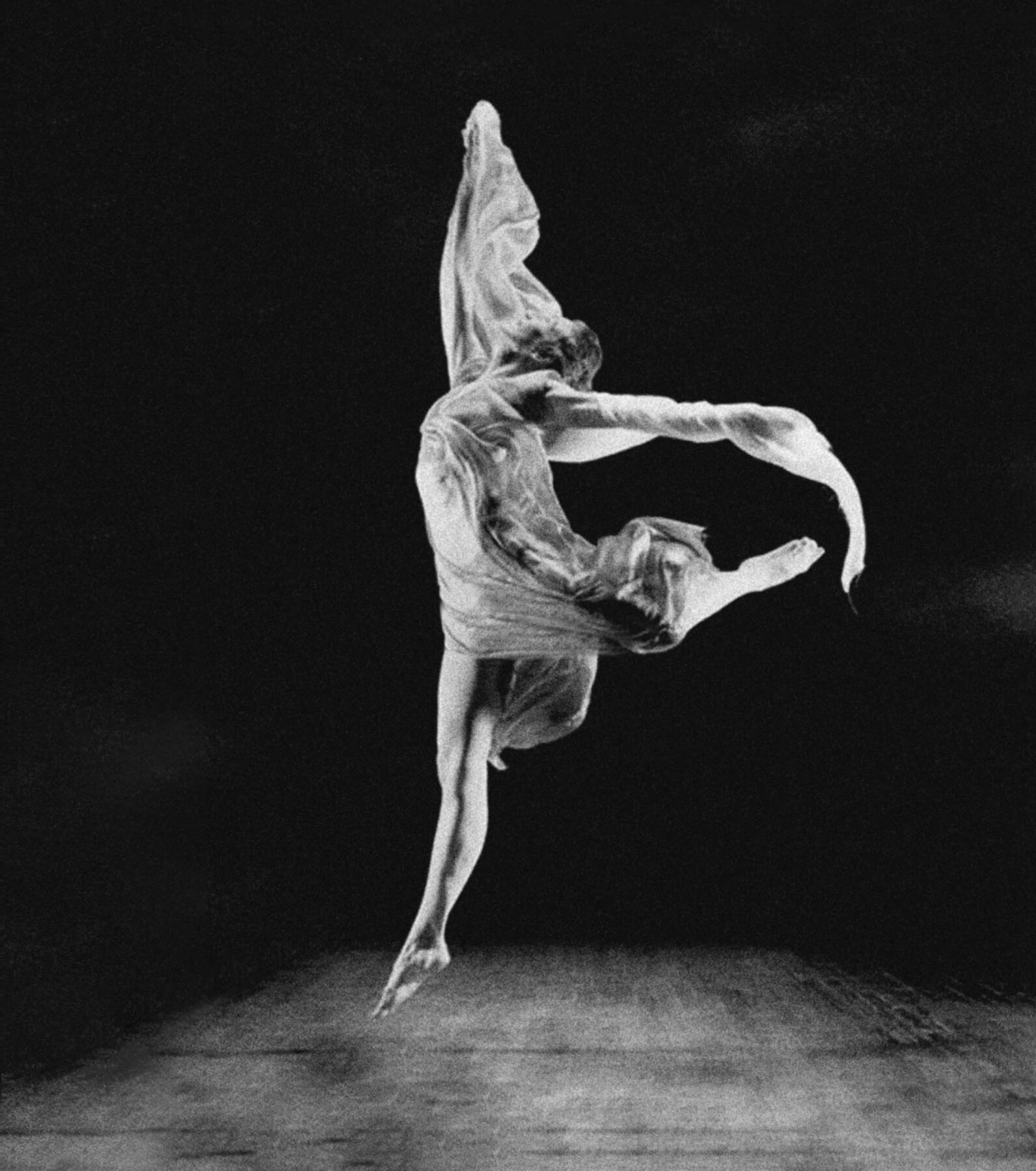 Fotografia em preto e branco. Mulher em movimento de dança no palco. Ela tem cabelo médio preso, usa um vestido esvoaçante e está descalça, saltando no ar, com uma das pernas dobrada para trás, os braços abertos e a cabeça inclinada para trás.