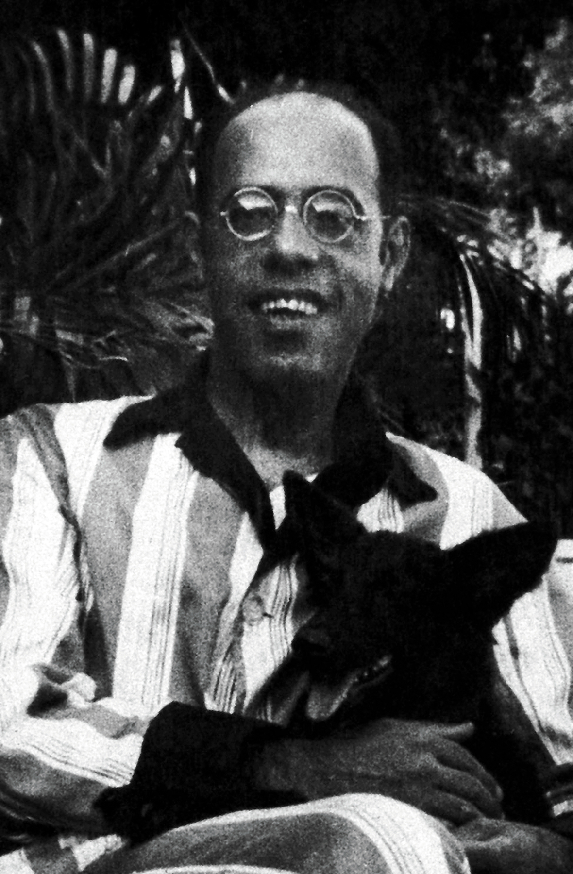 Fotografia em preto e branco. Homem calvo e de óculos, vestido com uma camisa com listas verticais. Ele sorri e segura no colo um cachorro preto de orelhas grandes.