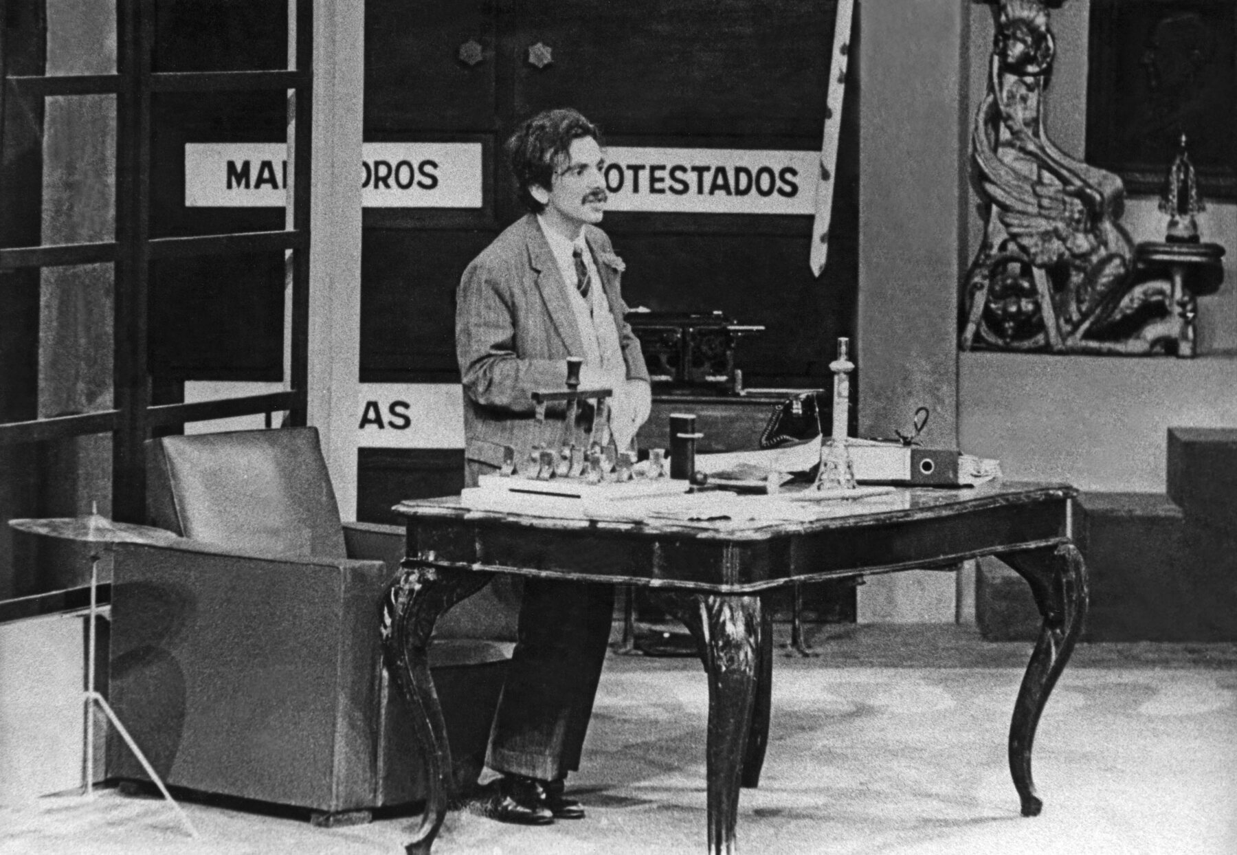 Fotografia em preto e branco. Homem de cabelo curto e bigode, vestido com paletó e sapato social, está em pé, entre uma poltrona e uma mesa antiga. Ao fundo, observa-se alguns letreiros nas portas.