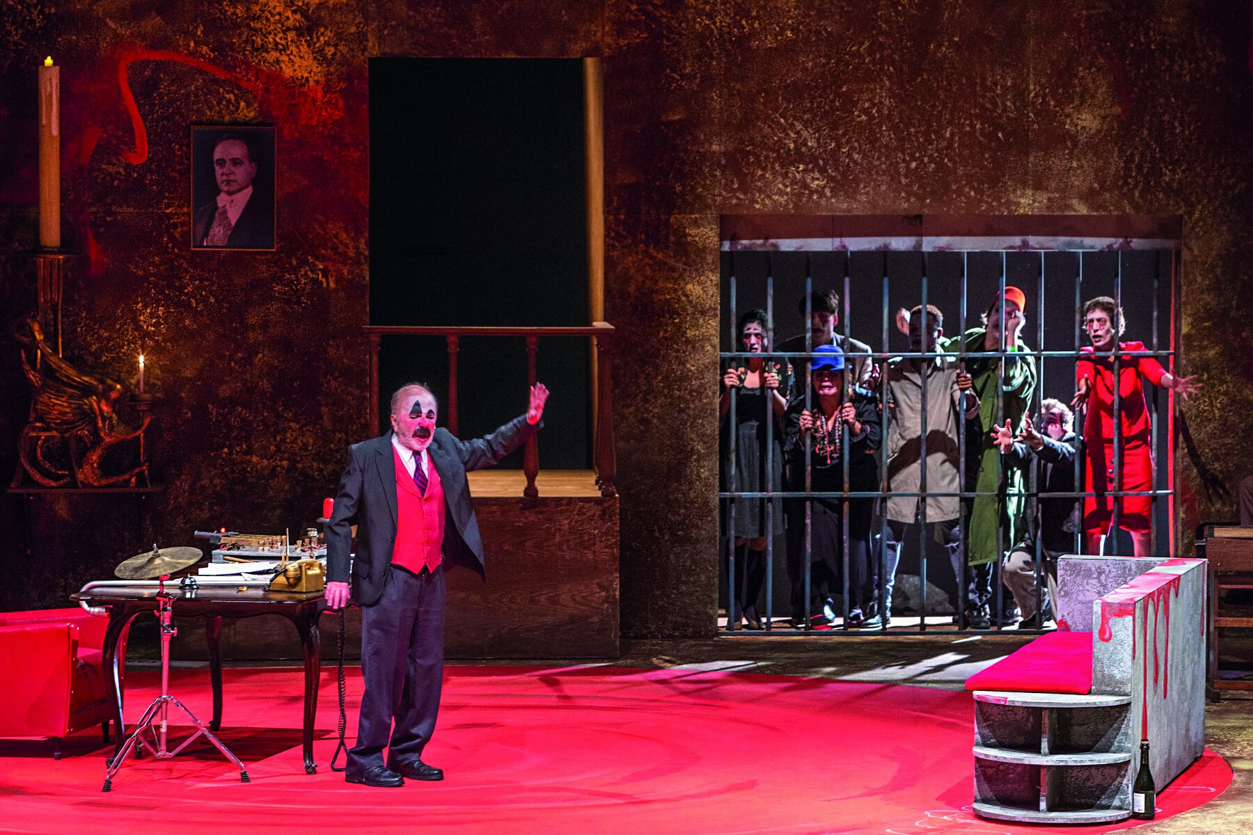 Fotografia. Apresentação teatral. Homem idoso com o rosto pintado imitando um grande bigode preto e uma mancha no olho, vestido com paletó cinza e colete vermelho. Ele está à frente de uma mesa, em um cenário que remete a um escritório. Ao fundo, do lado esquerdo, há uma fotografia do ex-presidente Getúlio Vargas na parede. À direita, pessoas se aglomeram em uma cela de prisão. O ambiente tem tons em vermelho, incluindo o chão.