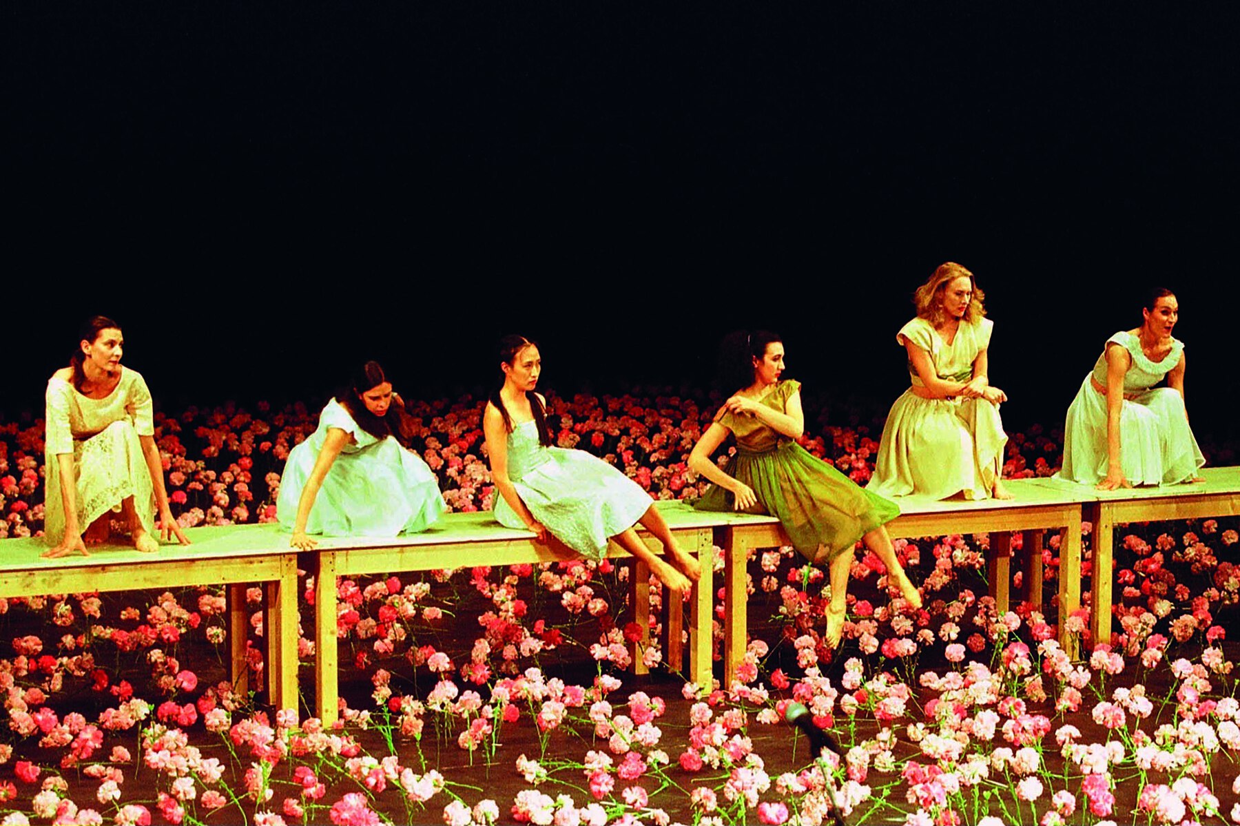 Fotografia. Seis mulheres usam vestido em tons de azul e verde e estão sentadas em mesas de madeira dispostas lado a lado sobre um palco florido. O chão está coberto de flores cor-de-rosa.
