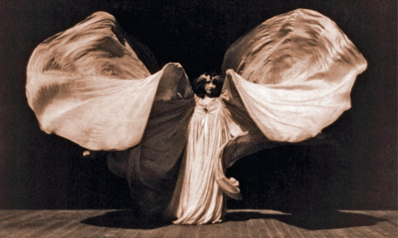 Fotografia em preto e branco. Dançarina no palco, vestida com uma roupa clara esvoaçante. Os braços estão em movimento e simulam asas com o tecido solto do vestido.