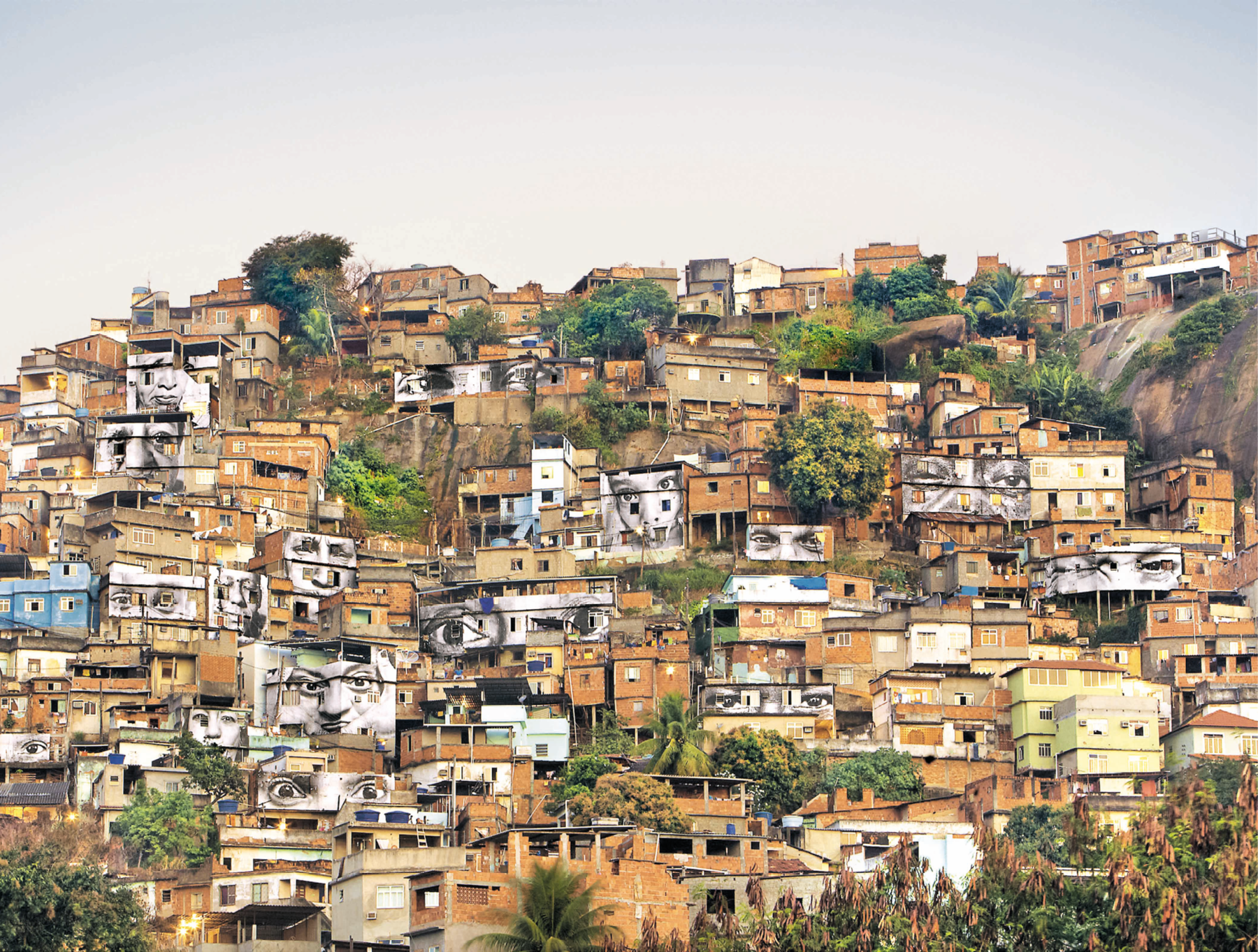 Fotografia. Vista geral de um morro totalmente ocupado por casas populares aglomeradas. Há pinturas de rostos humanos em preto e branco nos muros de algumas casas. O conjunto dessas pinturas compõem a intervenção artística.