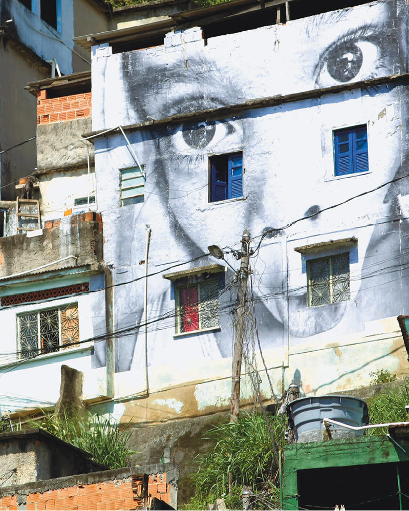 Fotografia. Vista de um muro lateral de uma casa de dois andares com grafite de um rosto de uma mulher por todo o muro.