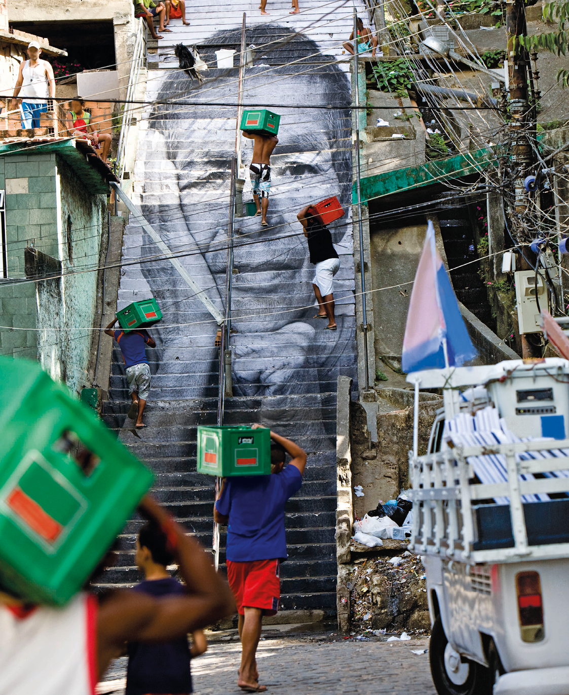 Fotografia. Em um cenário com casas com os tijolos aparentes, uma grande escadaria e homens transportando caixas verdes. Há também um automóvel perua, postes com muitos fios elétricos e uma bandeira.
Na escadaria, vê-se, em tamanho grande, um grafite em preto e branco com o rosto de uma mulher.