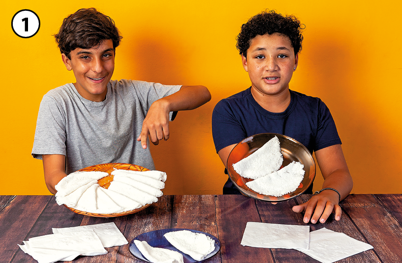 Fotografia 1: Dois garotos vestindo camisetas estão atrás de uma mesa de madeira. Eles mostram para o observador dois pratos com tapiocas. Há outras tapiocas dispostas sobre a mesa.