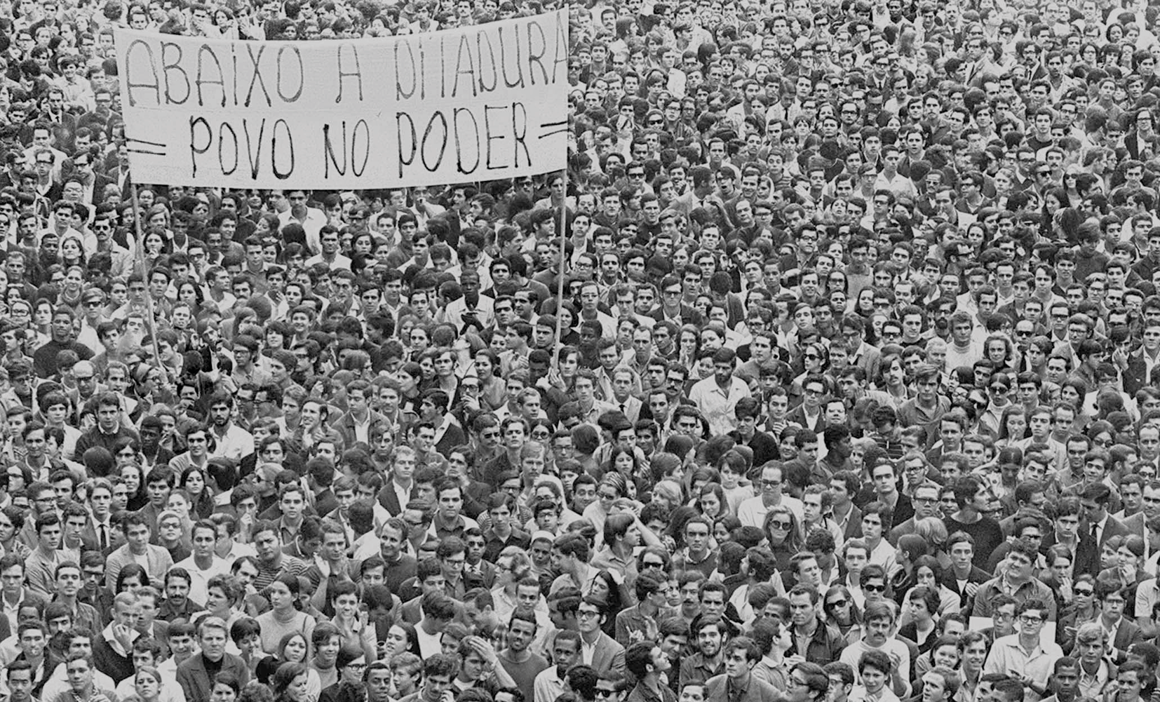 Fotografia em preto e branco. Uma multidão de pessoas reunidas carregando uma grande faixa com os dizeres: Abaixo a ditadura, povo no poder.