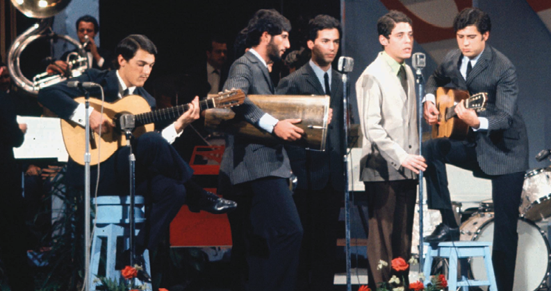 Fotografia. Grupo de músicos vestidos de terno cantam e tocam instrumentos musicais, como violões, tuba e tambor. À frente deles, microfones de chão.