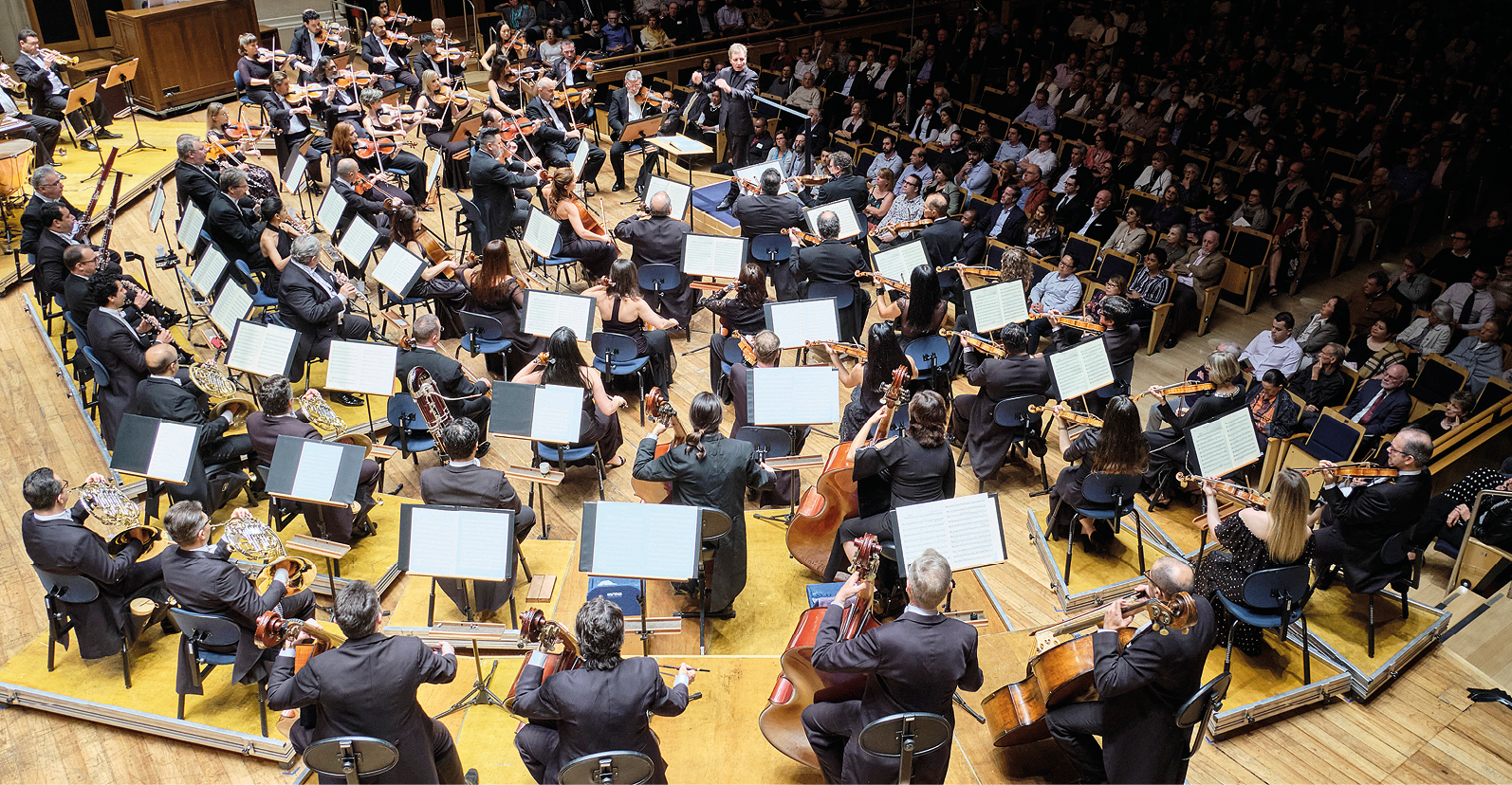 Fotografia. Vista superior de parte de uma orquestra em um palco formando um semicírculo. Destaque para região onde pessoas tocam instrumentos de cordas.