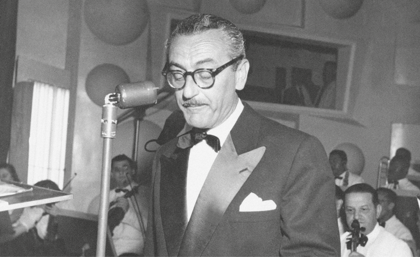 Fotografia em preto e branco. Homem de cabelo curto e óculos de armação arredondada, vestindo camisa, gravata e paletó. Está em frente a um microfone.