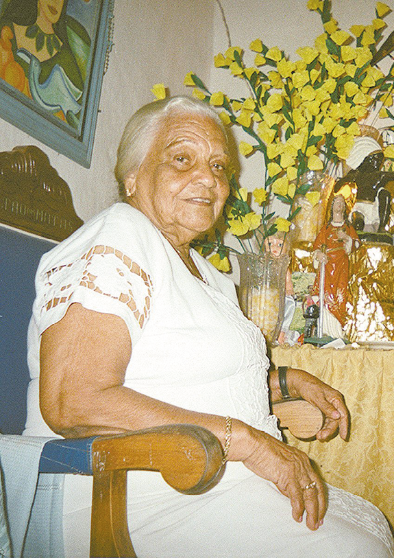 Fotografia. Mulher idosa de cabelo grisalho, com vestido branco rendado nas mangas, sentada em uma cadeira de madeira. Ao fundo, detalhe de um quadro na parede à esquerda e, à direita, uma mesa com vaso de flores amarelas, um copo de vela e uma imagem de santa.