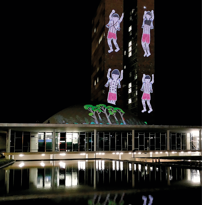 Fotografia. Projeção luminosa na lateral de um prédio, na qual se veem quatro pessoas indígenas e árvores. A fotografia foi tirada à noite. Há um espelho-d'água à frente que reflete a projeção.