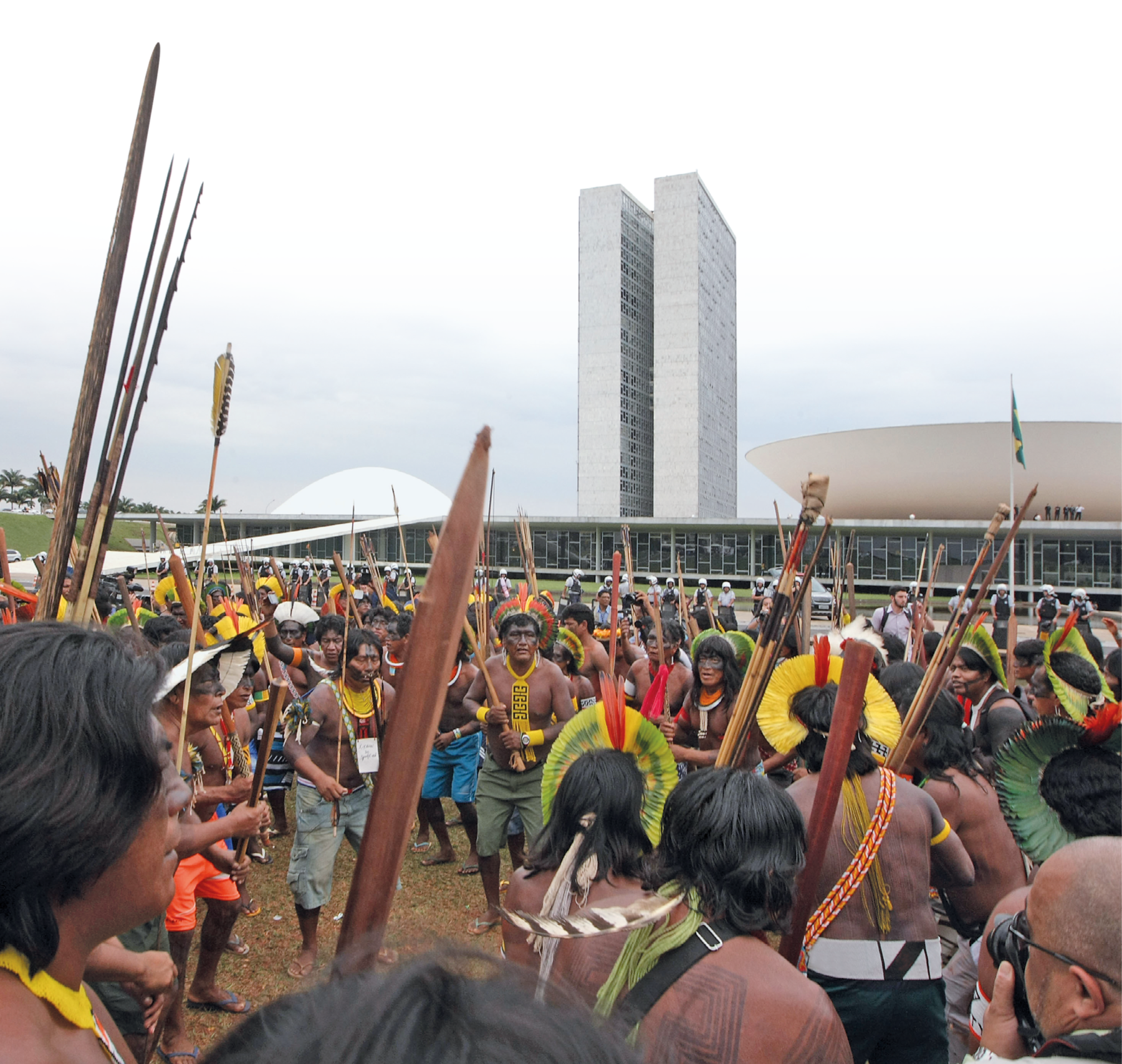 Fotografia. Homens indígenas em manifestação em frente ao Congresso Nacional, em Brasília. Muitos usam cocar e colar e estão com o rosto e o corpo pintados, carregando arcos e flechas, além de bastões de madeira. Ao fundo, há policiais acompanhando a manifestação de longe e os prédios do Congresso Nacional.