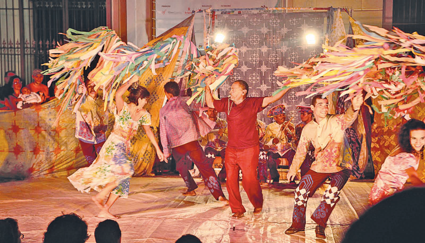 Fotografia. Grupo de dança com homens e mulheres vestidos com roupas de cores vibrantes. Eles movimentam para o alto fitas e bandeiras coloridas. Ao centro, homem com roupa vermelha traz um apito na boca. Ao fundo, músicos sentados tocam instrumentos musicais.