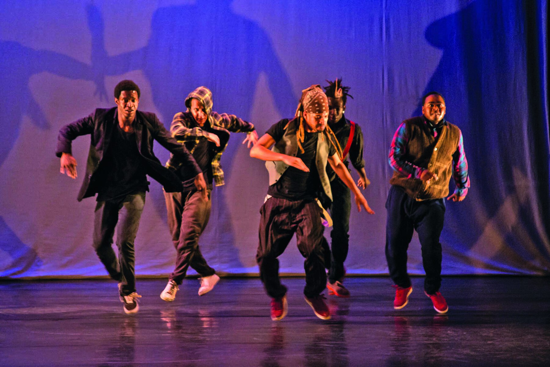 Fotografia. Palco com cinco homens de roupas em tons de preto, marrom e cinza. Estão em movimento de dança. Ao fundo, sombra dos dançarinos projetada em uma cortina branca.