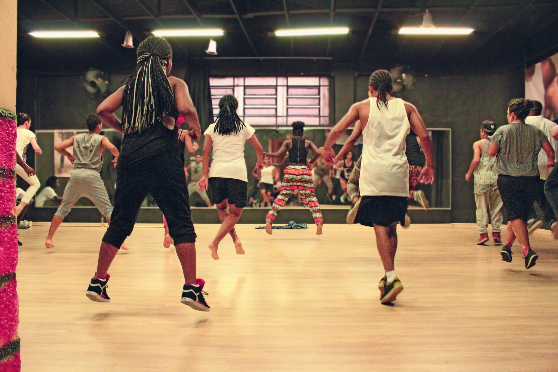 Fotografia. Interior de um salão em que algumas pessoas estão fazendo aula de dança. O grupo é formado por homens e mulheres e todos usam roupas esportivas, como bermuda e regata. Alguns estão calçados, outros estão descalços. Estão de costas. Ao fundo, um grande espelho horizontal disposto ao longo da extensão da parede. Ele reflete parte do grupo.