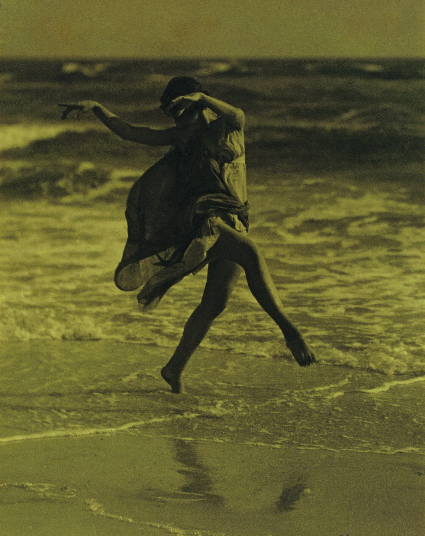 Fotografia em tons de sépia. Mulher de cabelo curto, vestido médio esvoaçante, dança na beira-mar.