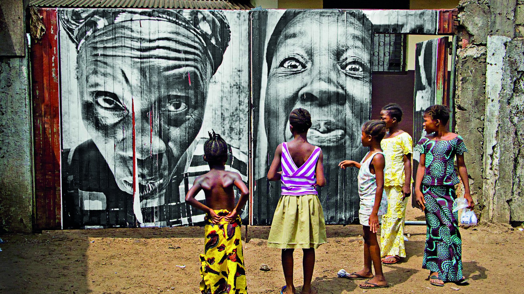 Fotografia. Em primeiro plano, cinco crianças vestidas com roupas estampadas coloridas estão em pé, de costas, observando um muro com intervenção artística. Ao fundo, grafite realista em preto e branco de rostos de duas mulheres com turbantes na cabeça.