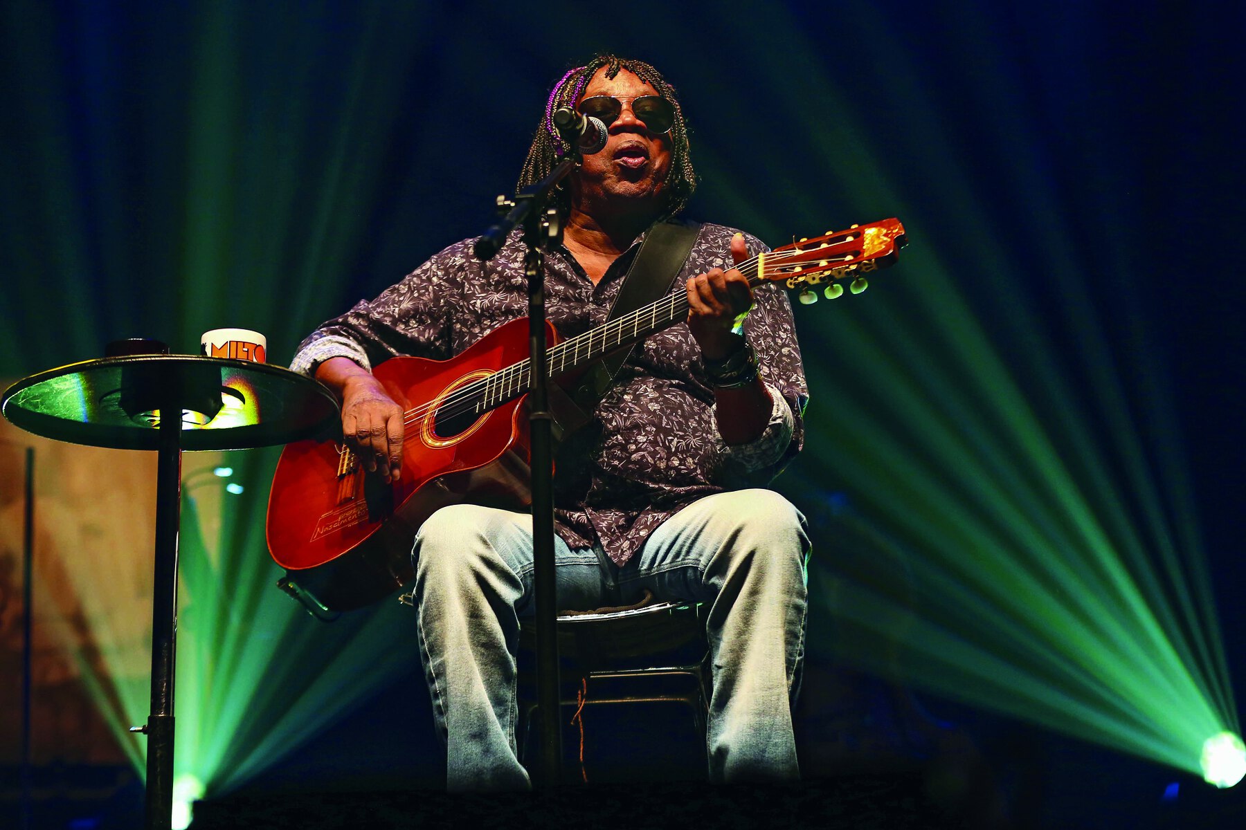 Fotografia. Homem sentado em um banquinho toca violão em um palco. Ele tem cabelo escuro médio trançado e usa óculos escuros, camisa estampada marrom com detalhes em branco e calça clara. À esquerda, uma mesinha de apoio e um holofote que projeta luz do chão. À direita, outro holofote.