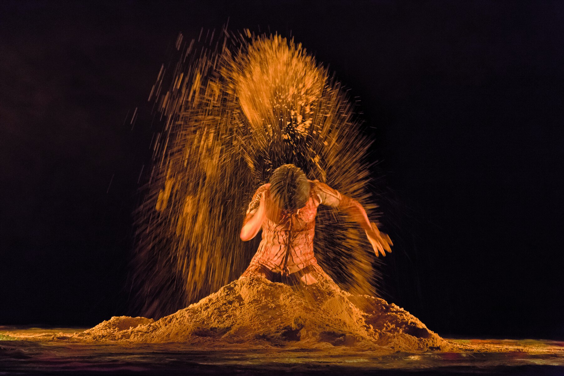 Fotografias. Duas fotografias mostram uma mulher em movimento de dança em um palco com um monte de terra. Ela veste roupa justa em tons de marrom. Na primeira imagem, ela está agachada, com a cabeça e o tronco inclinados para baixo,  e joga a terra por cima do corpo; na segunda imagem, também agachada, ela joga a terra à frente dela.