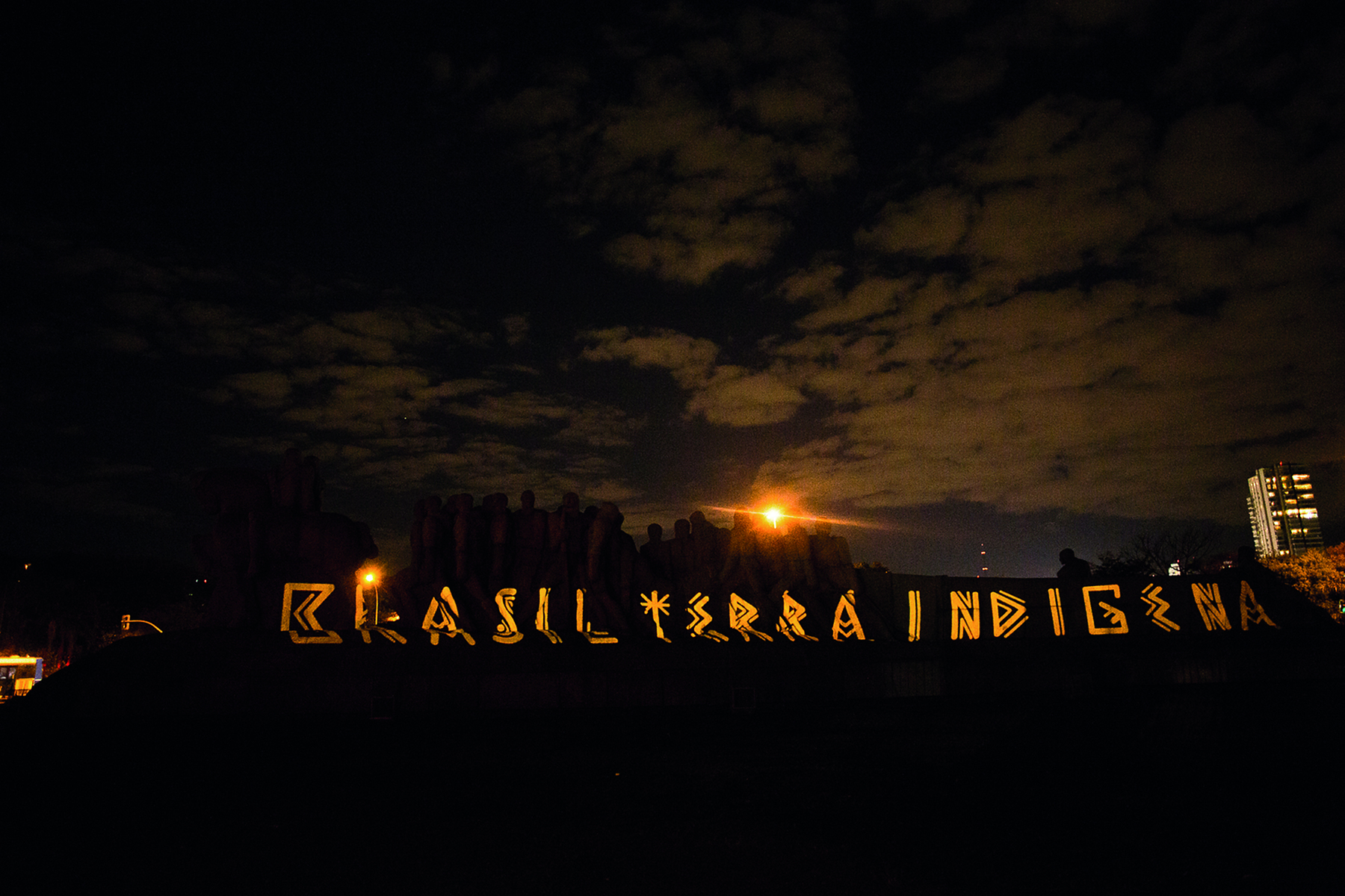 Fotografia. Projeção da frase Brasil Terra Indígena em um monumento ao entardecer. As letras apresentam grafismo que remete à arte indígena. Céu com nuvens.