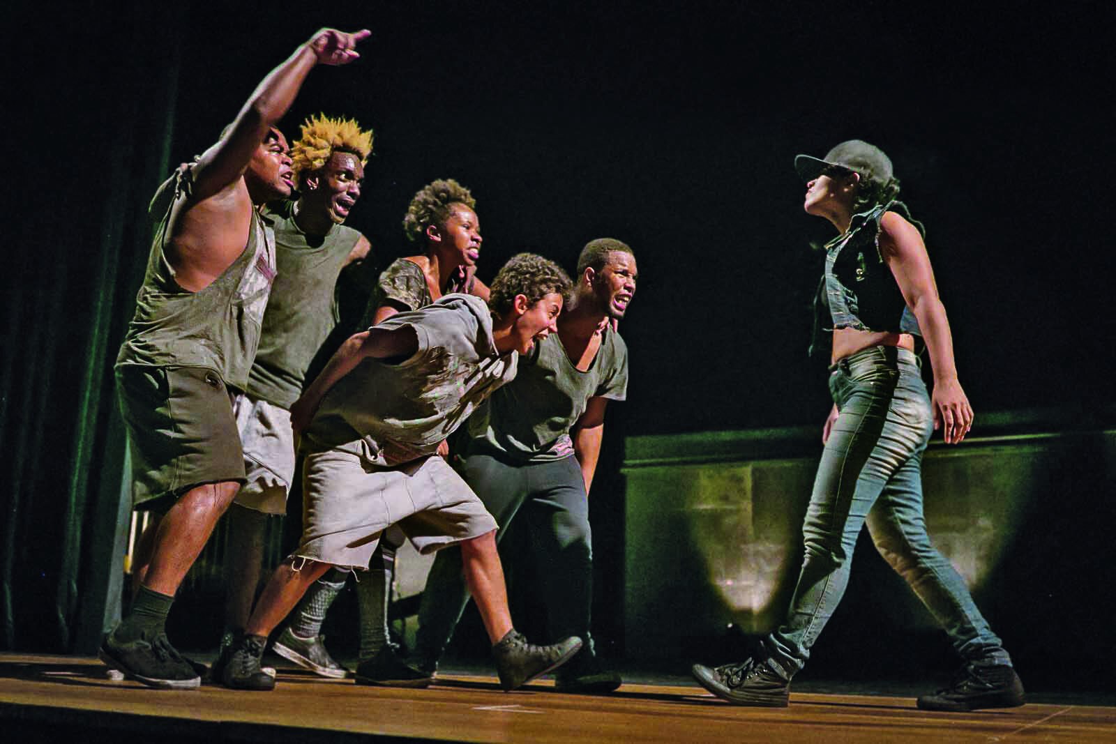 Fotografia. Grupo de dançarinos no palco. À esquerda, um grupo de cinco pessoas confronta uma mulher, que está à direita da foto. Todos usam roupas informais em tons de cinza e estão em movimento de dança.