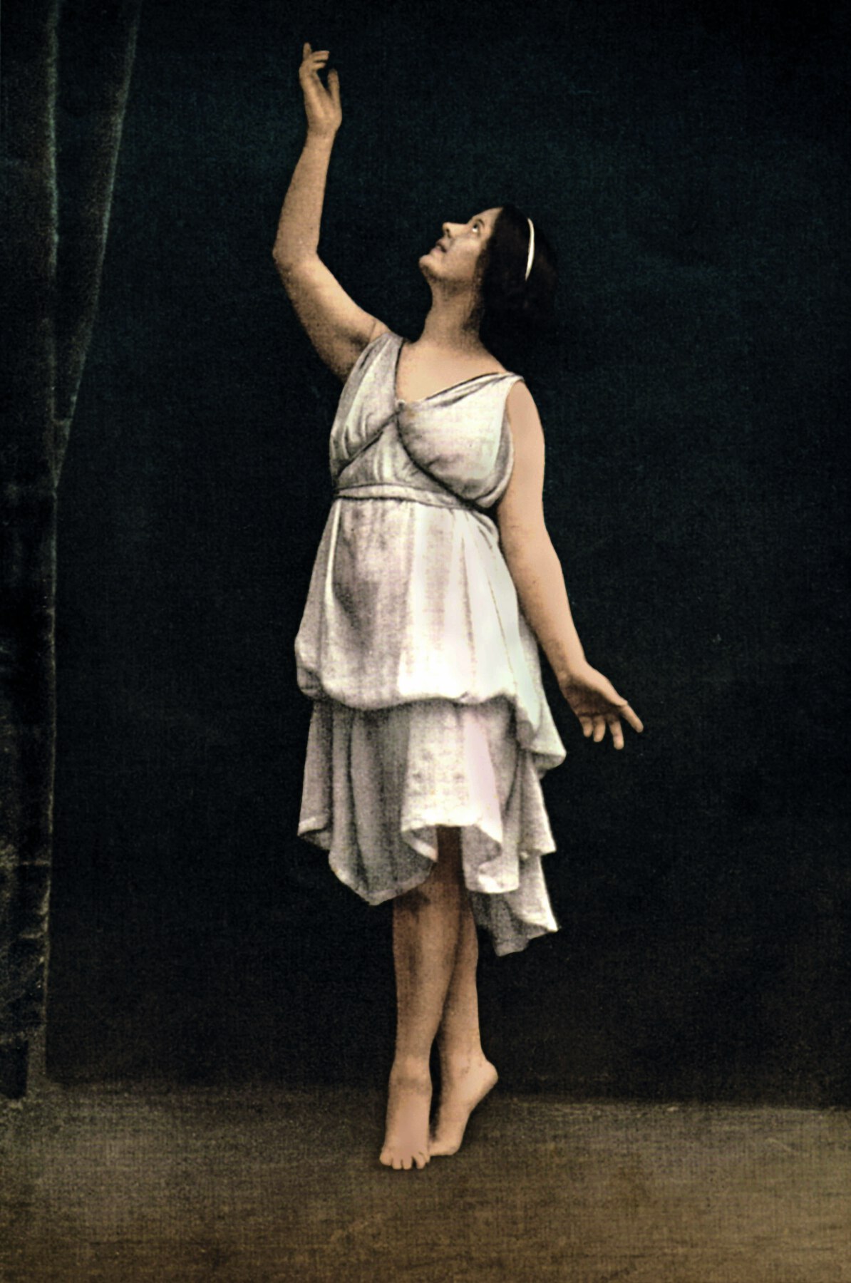 Fotografia. Mulher dança em um palco. Ela tem cabelo preto e usa um vestido claro médio. Está descalça na ponta dos pés, com o braço direito erguido e o rosto voltado para a mão direita. A mão esquerda permanece ao lado do corpo.