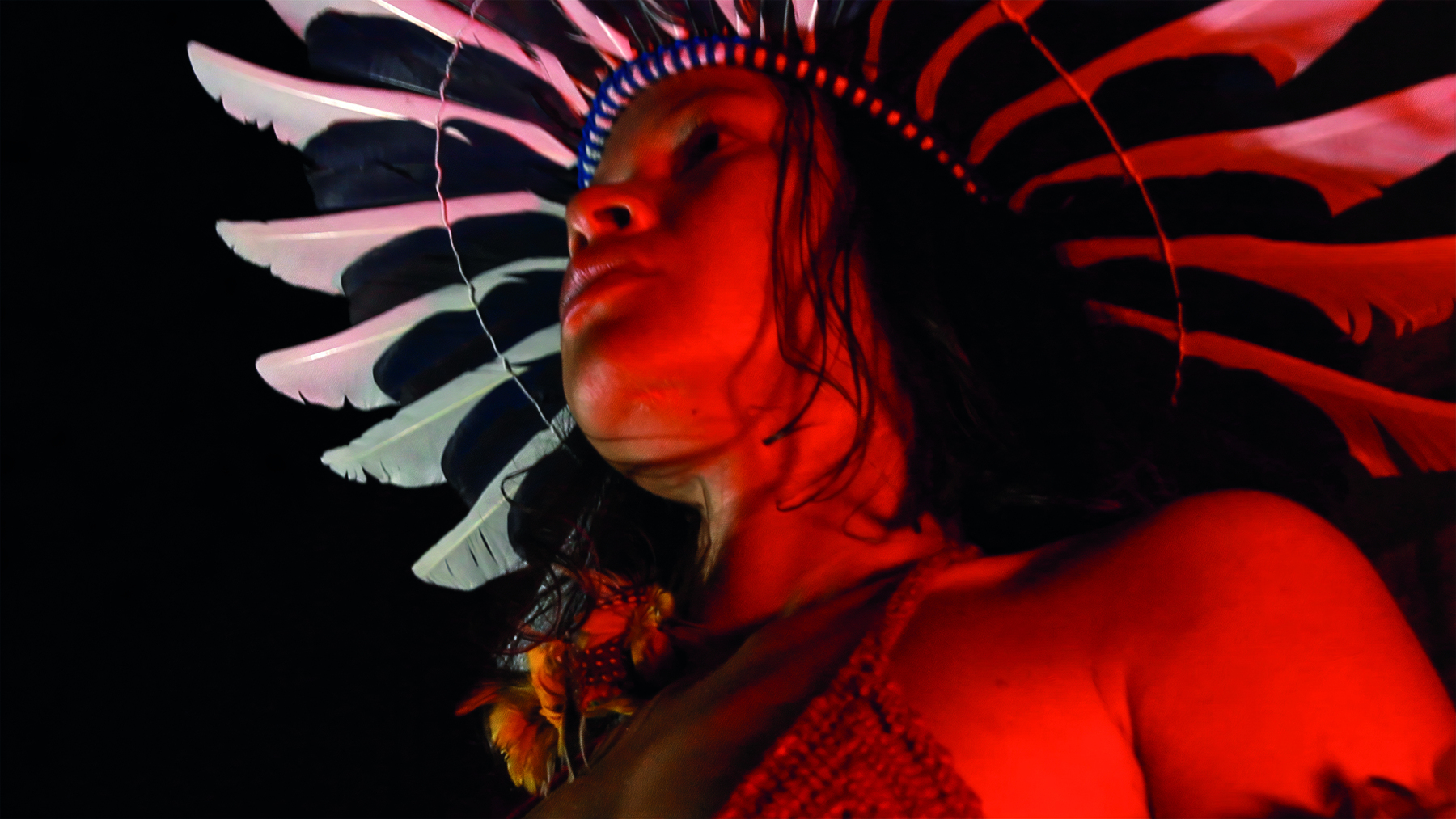 Fotografia. Destaque do rosto de uma mulher indígena com cocar de penas claras. A imagem tem tons avermelhados.