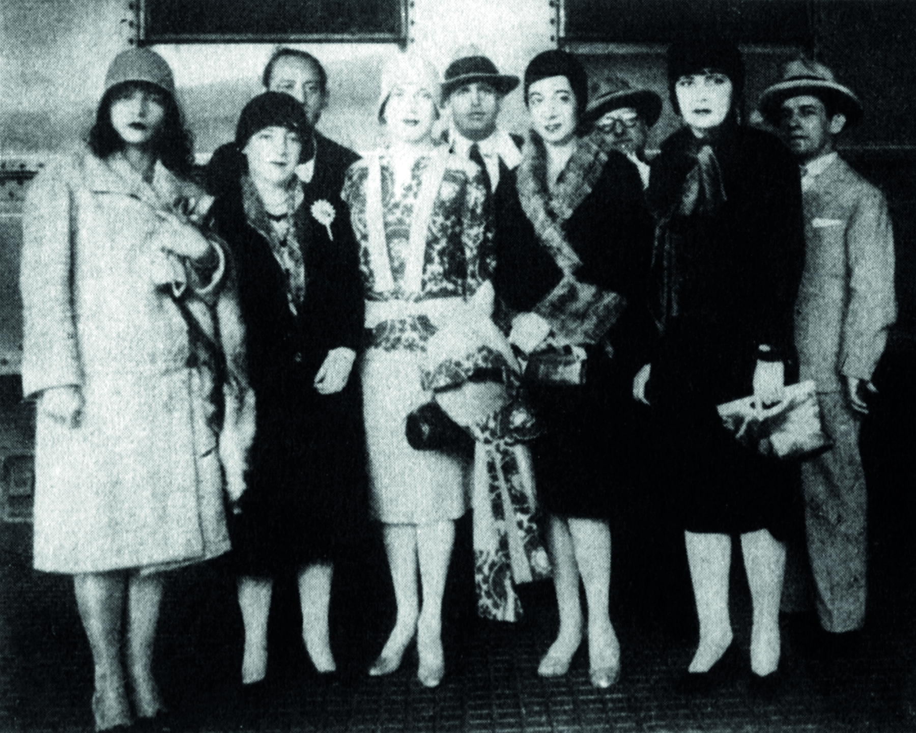 Fotografia em preto e branco. À frente, cinco mulheres vestidas elegantemente com chapéus pequenos, vestidos e casacos. Atrás, quatro homens de paletós e chapéus.