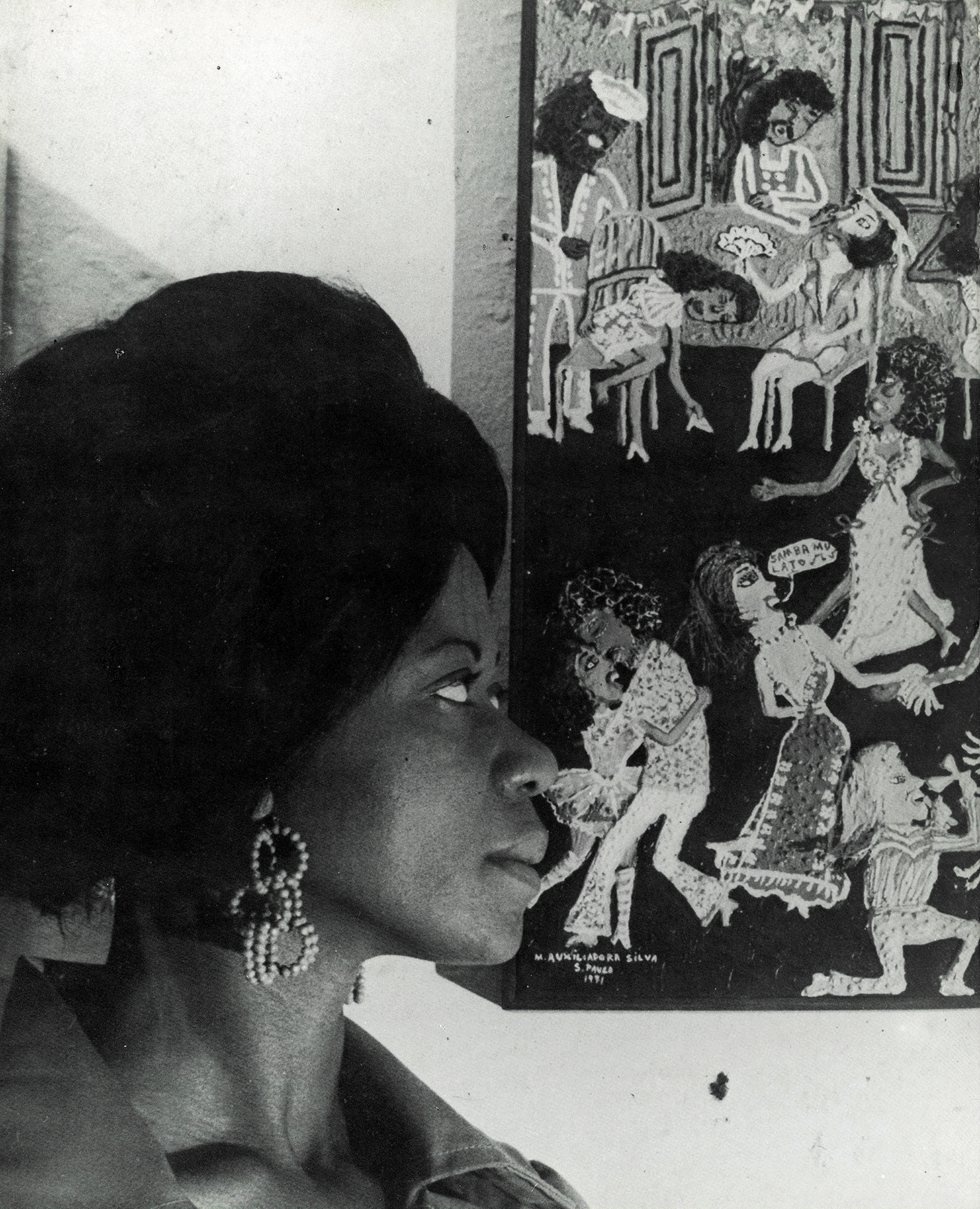 Fotografia em preto e branco. À esquerda, uma mulher de turbante e brincos grandes, de perfil, observa um quadro. À direita, um quadro que representa homens e mulheres dançando.