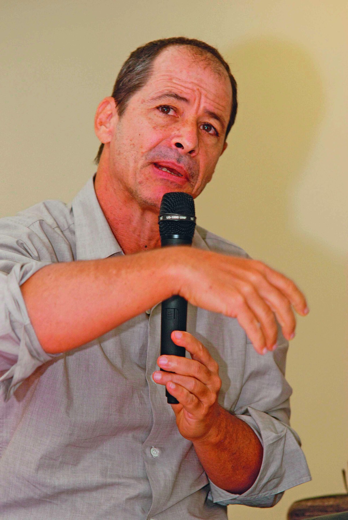 Fotografia. Homem de cabelo curto, calvo, veste uma camisa cinza e fala segurando um microfone na mão.