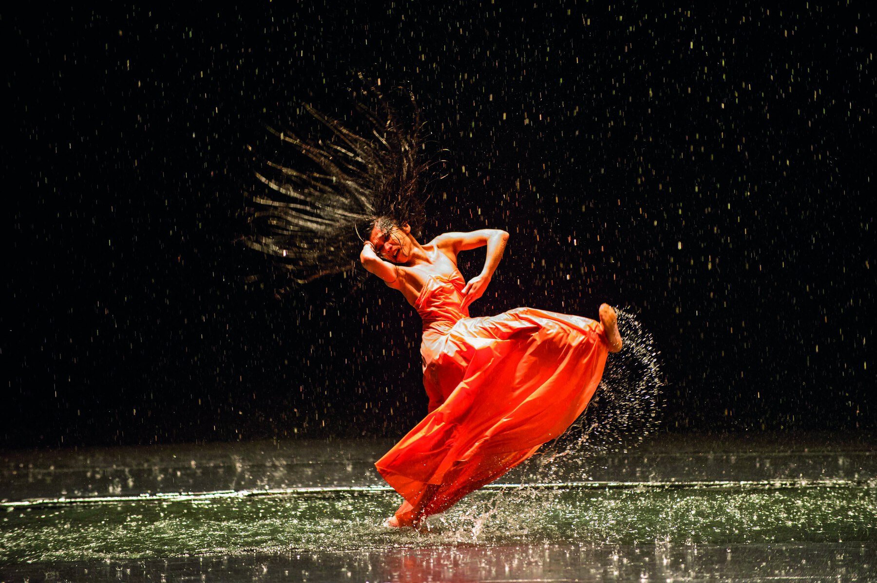 Fotografia. Imagem impactante de mulher dançando com água. Ela tem cabelo comprido escuro e usa um vestido longo laranja. Sua perna direita está levantada, respingando água, o tronco virado para a direita, com o braço direito atrás da cabeça e o esquerdo dobrado à frente do corpo. O chão tem uma poça de água grande e o fundo é escuro.