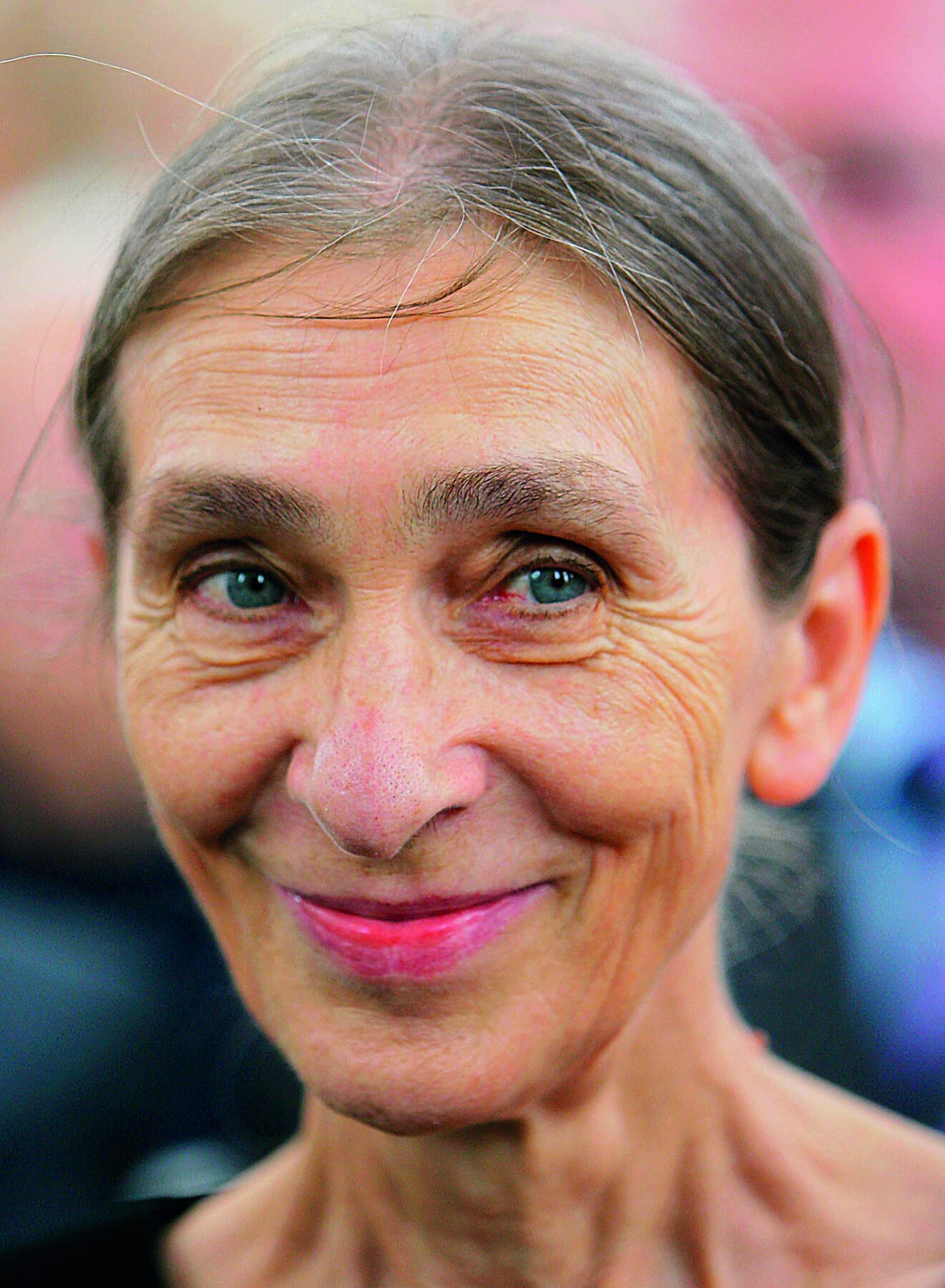 Fotografia. Retrato de uma mulher idosa de cabelo grisalho preso. Ela sorri.