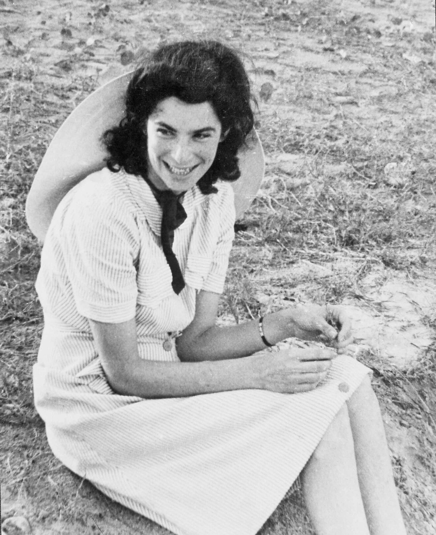 Fotografia em preto e branco. Mulher sentada no chão gramado de um espaço ao ar livre. Ela tem cabelo escuro médio, usa um vestido claro e está sorrindo. Usa um chapéu claro apoiado nas costas.