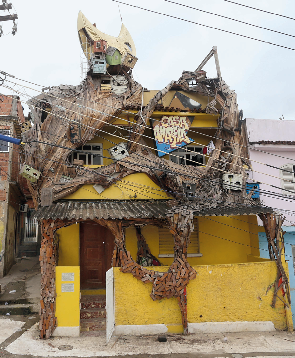 Fotografia. Fachada de uma casa amarela de dois andares. Encobrindo o topo da casa, uma intervenção artística criada a partir de uma estrutura de madeira que simula a raiz de uma árvore, na qual se veem pequenas réplicas de casas da comunidade.