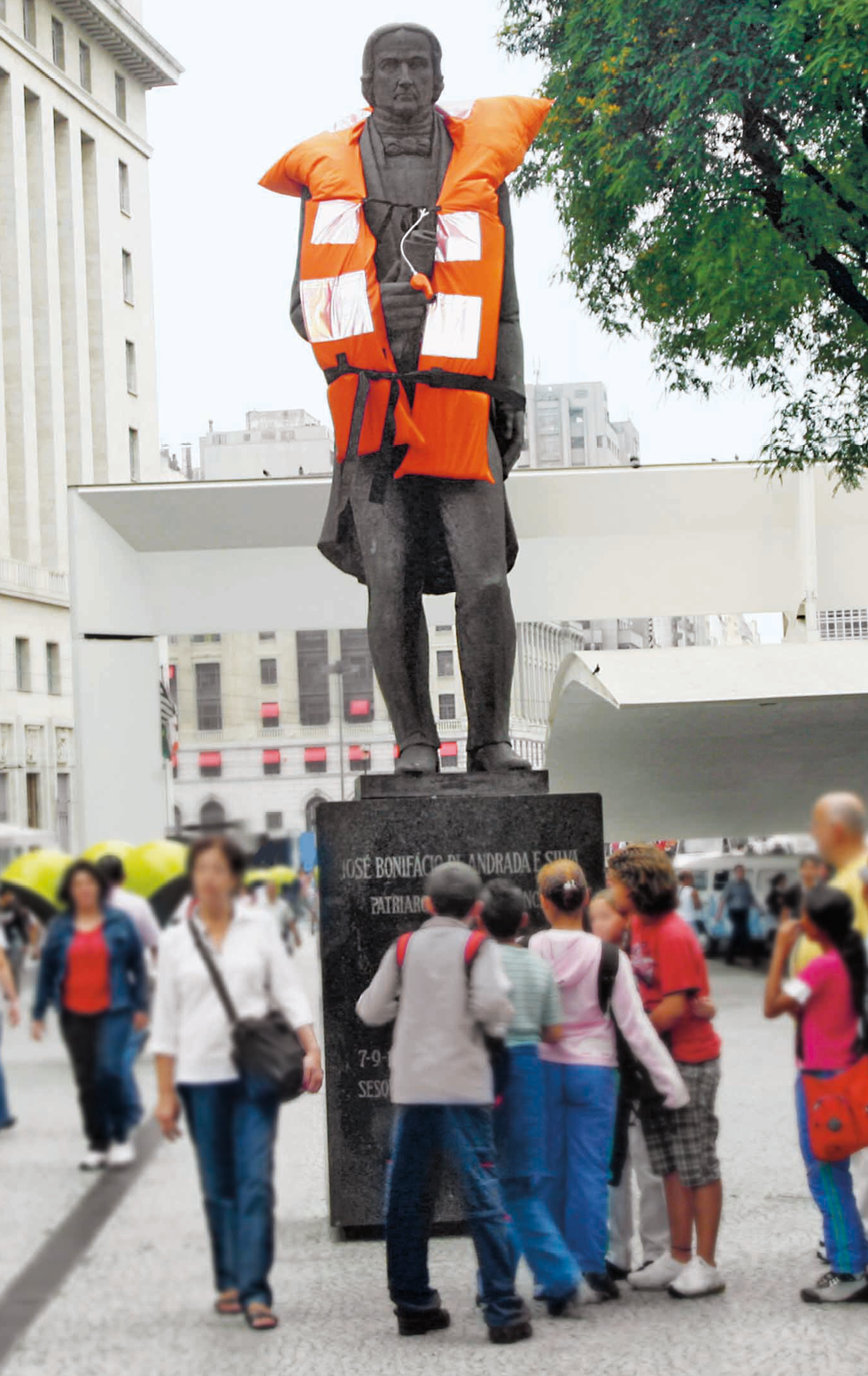 Fotografia. Estátua de homem vestido com um colete salva-vidas laranja real, posicionada ao centro de uma praça e sobre um pedestal com texto. Algumas pessoas estão bem próximas à estátua, observando o texto do pedestal. À esquerda e ao fundo, construções e pessoas caminhando.