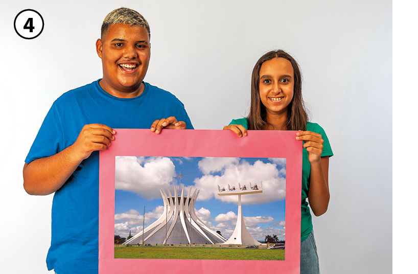 Fotografia. 4: Dois jovens segurando um cartaz. À esquerda, menino de cabelo curto descolorido vestido com uma camiseta azul. Ao lado, menina de cabelo longo castanho, vestida com camiseta verde. Eles estão segurando um cartaz com a fotografia de um monumento arquitetônico de linhas modernas, que lembra a coroa de um abacaxi.