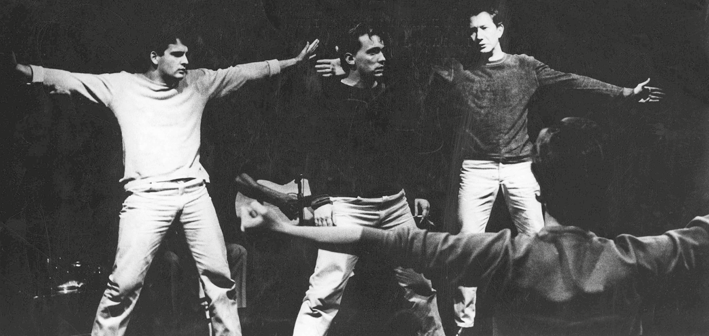 Fotografia em preto e branco. Quatro atores encenam uma peça teatral. Eles estão vestidos com calças jeans e camisetas de manga longa e estão de braços abertos no palco, um deles de costas.