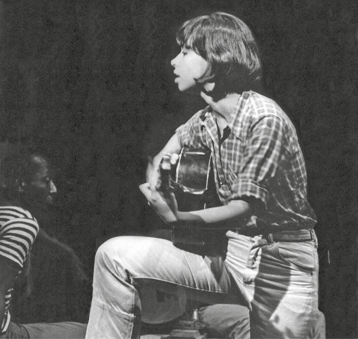 Fotografia em preto e branco. Perfil de cantora tocando violão. Ela está apoiando o violão na perna dobrada um pouco acima do nível do solo. Tem cabelo escuro liso e veste camiseta xadrez e calça.