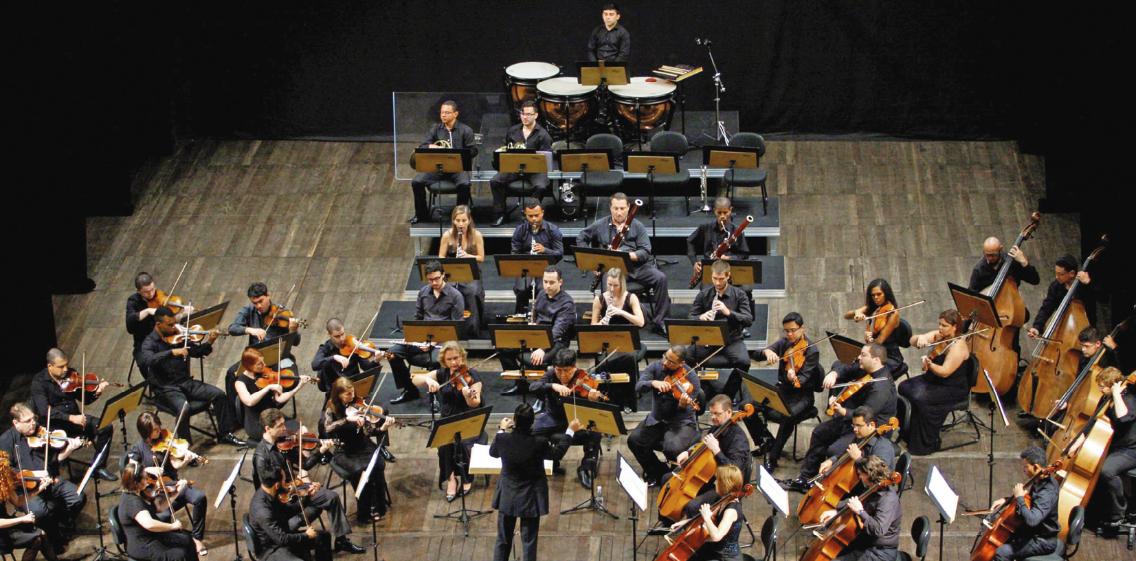 Fotografia. Vista de cima de uma orquestra posicionada no palco. No centro, o  maestro está em pé, de frente para a orquestra. À esquerda e à direita, estão músicos tocando instrumentos de corda. No centro, ao fundo, estão músicos tocando instrumentos de sopro e de percussão.