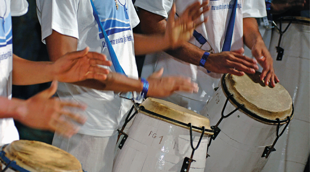 Fotografia. Destaque de mãos tocando atabaques. Os músicos usam roupas brancas e as camisetas tem um símbolo azul no meio.