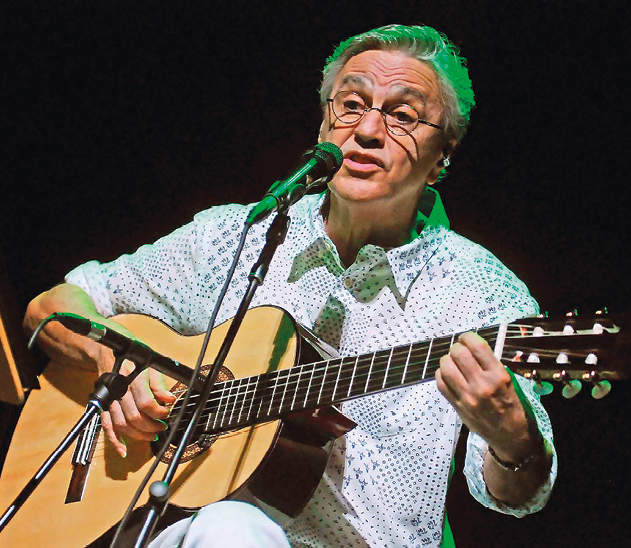 Fotografia. Homem de cabelo curto grisalho, óculos, com camisa branca com detalhes em azul-claro, toca violão e canta em um microfone.