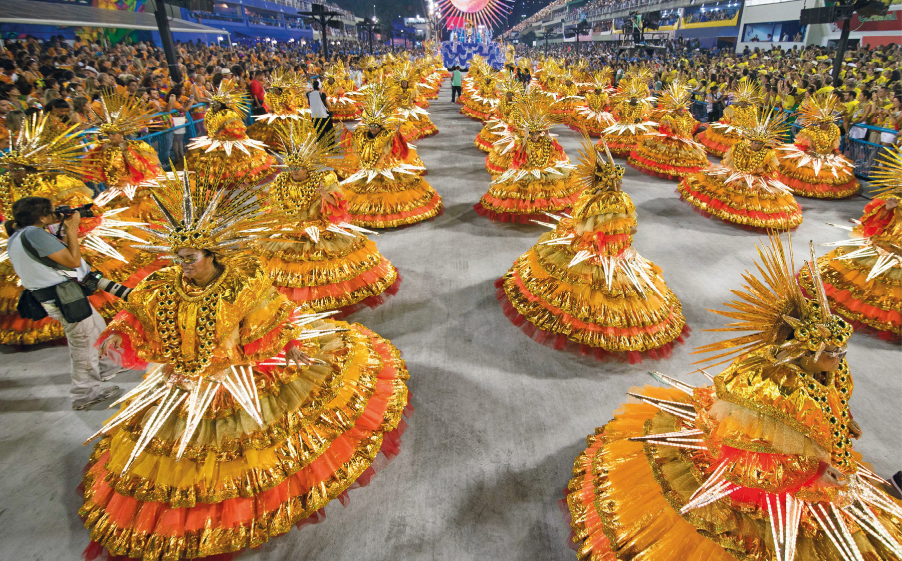 Fotografia. Vista de desfile de escola de samba. Em primeiro plano, mulheres vestidas de baianas, com fantasias luxuosas na cor laranja com detalhes dourados e plumagens. Ao fundo, o público nas arquibancadas. À esquerda, um fotógrafo está na ala documentando o evento.