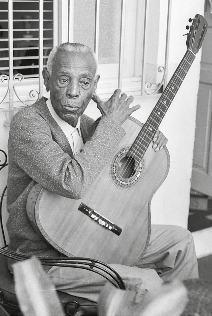 Fotografia em preto e branco. Homem idoso, com camisa clara e casaco, sentado em uma cadeira com um violão. Atrás, parte de uma janela com grades.