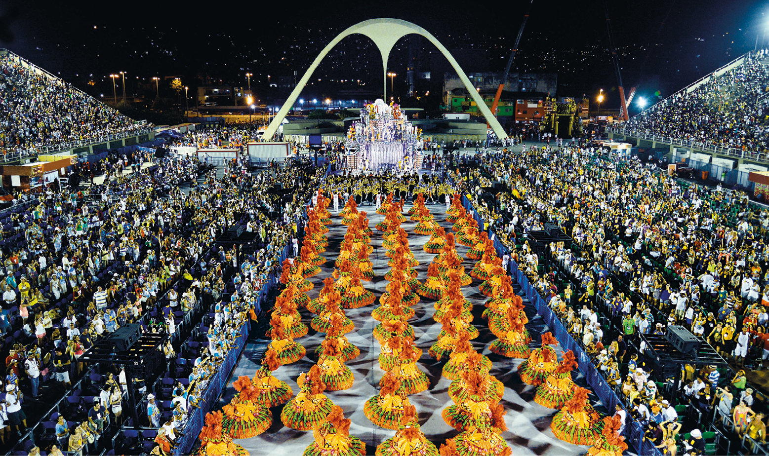 Fotografia. Vista de cima de um desfile de escola de samba. No centro da passarela, há mulheres vestidas de baianas, com roupas em tons de laranja e dourado. À esquerda e à direita, o público acompanha o desfile nas arquibancadas. Ao fundo, a praça da Apoteose com o arco de concreto.